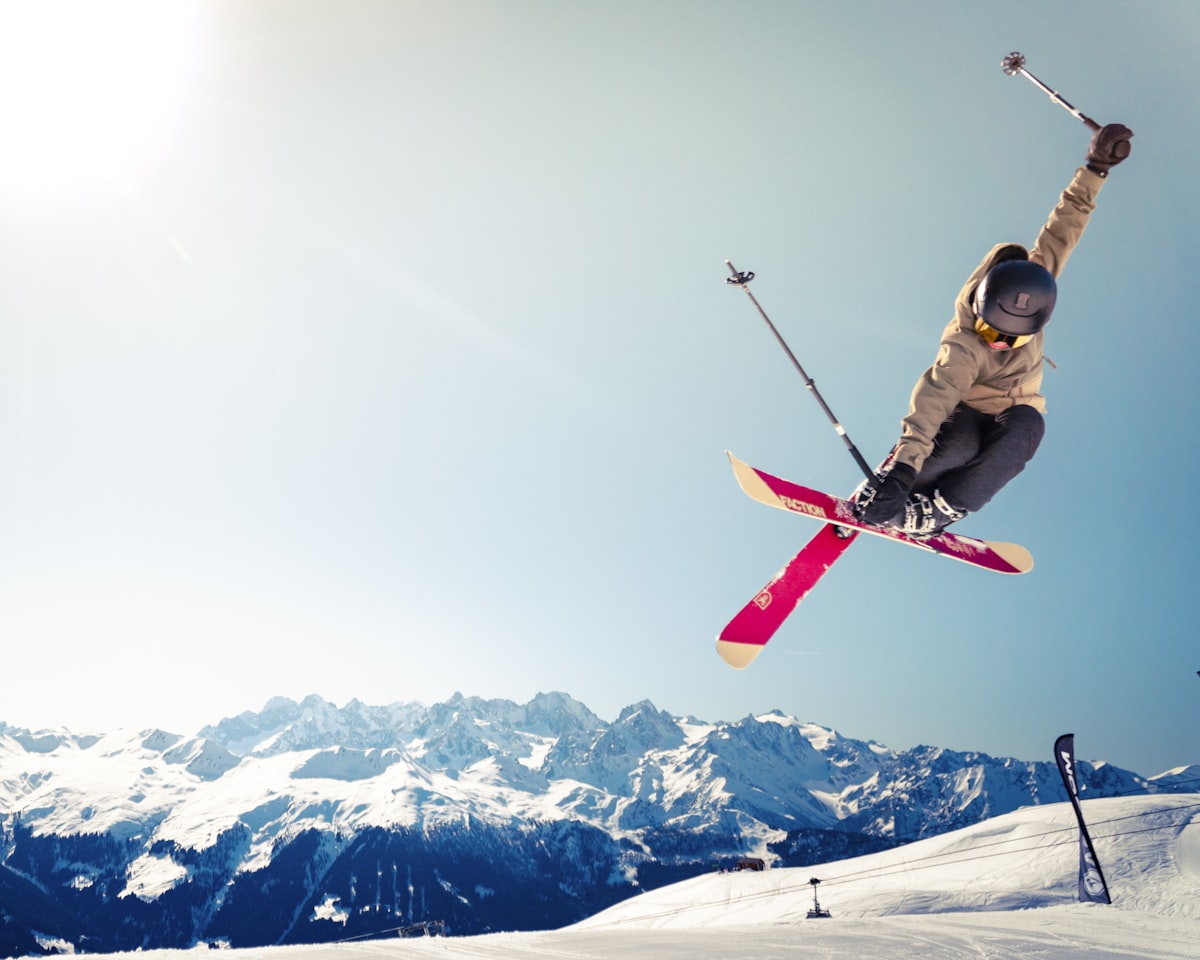 Skier launching off a jump against a backdrop of snow-covered alpine mountains and bright sunshine