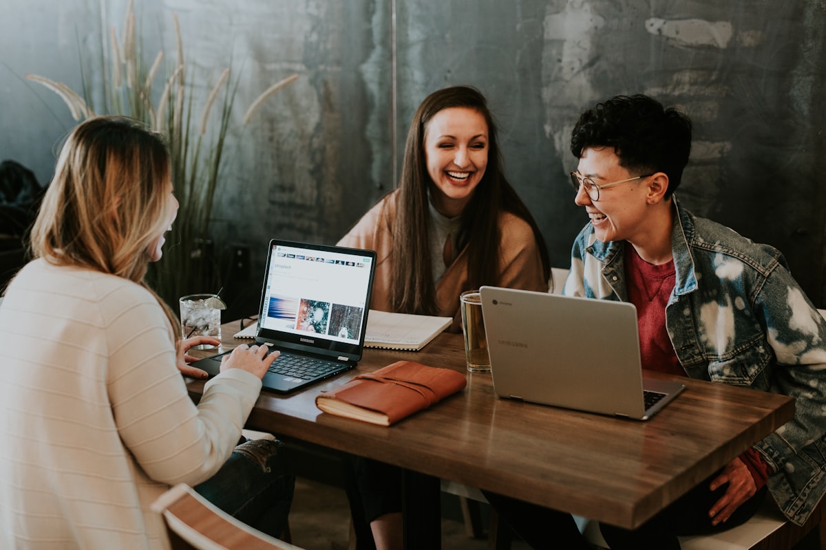 Three colleagues laughing and collaborating around laptops at a cafe table, capturing the energy of a small startup team