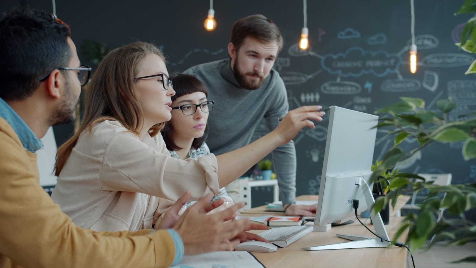 Colleagues gathered around a single laptop, one person pointing at the screen to teach the others